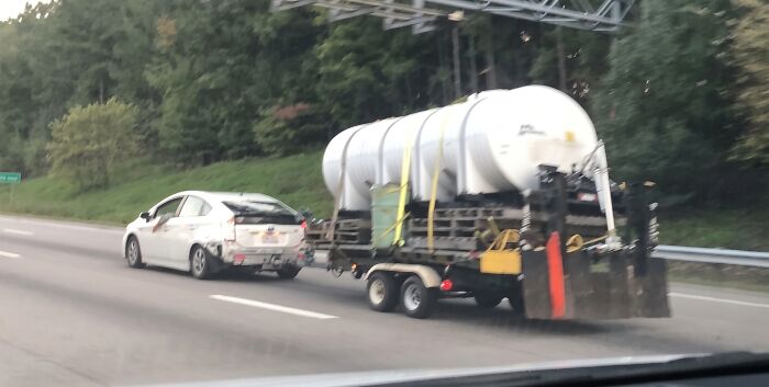 Small white car towing massive water tank trailer on highway in dangerous towing gone wrong situation