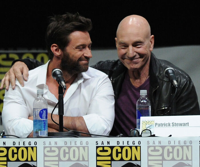 Hugh Jackman and Patrick Stewart sitting together smiling at a panel microphoned table. Hugh Jackman and Patrick Stewart sitting together smiling at a panel microphoned table.
