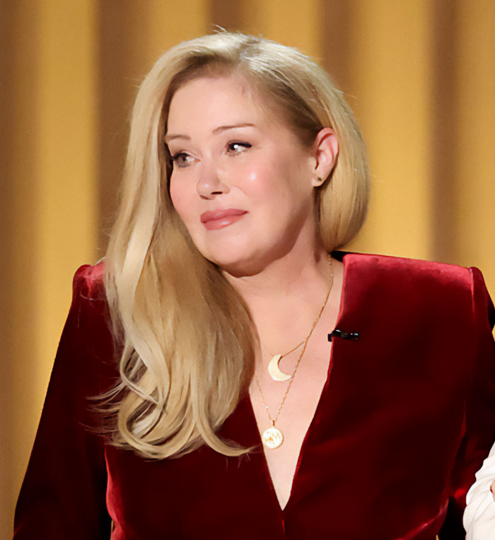 Christina Applegate wearing a red velvet top and gold necklaces while looking thoughtfully to the side.