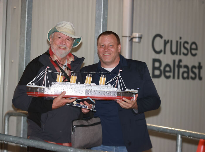 Two men holding a model ship near a permanent cruise terminal sign Two men holding a model ship near a permanent cruise terminal sign