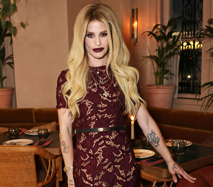 Kelly Osbourne standing in a dark burgundy lace dress with long blonde hair.