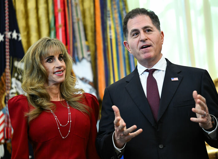 Woman in red dress standing beside man in dark suit speaking at official event Woman in red dress standing beside man in dark suit speaking at official event