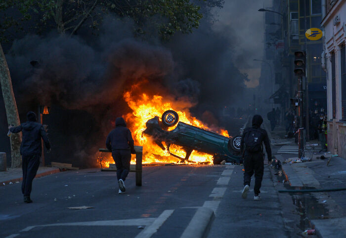 France riots scene with overturned car burning and people nearby on city street