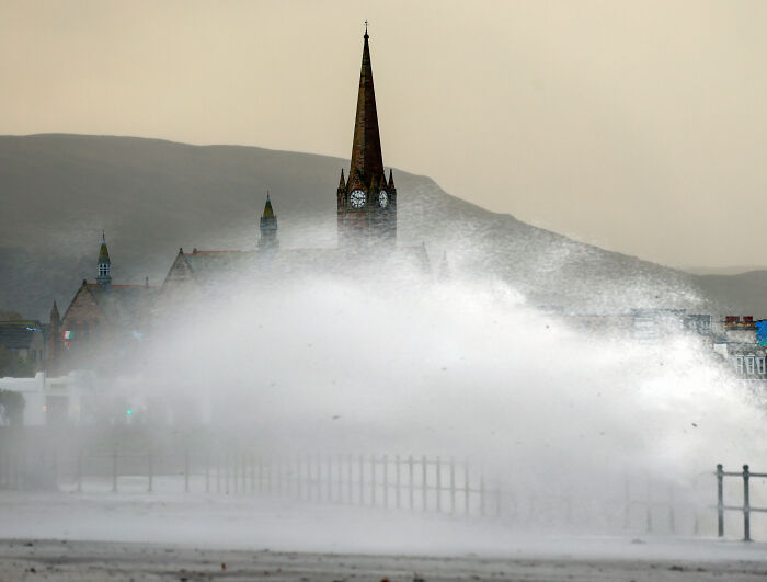 Large ocean wave crashing near a coastal church building causing severe weather flight disruptions.