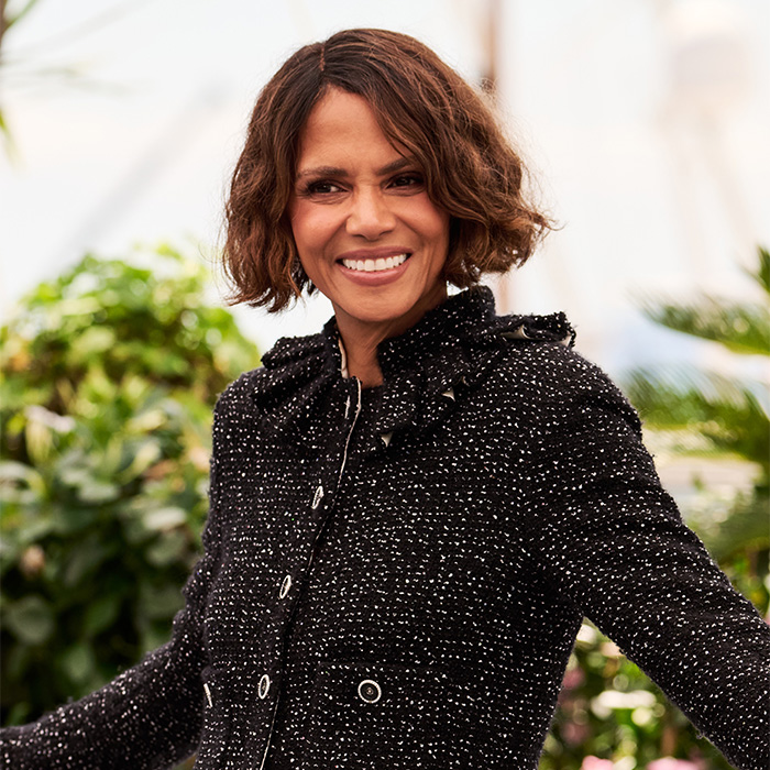 Hollywood actress Halle Berry smiling in a black and white tweed jacket with green foliage behind her.