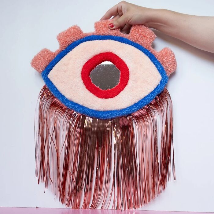 Tufted eye art piece with a mirror iris and pink tinsel fringe, held by artist Ina Dyreborg