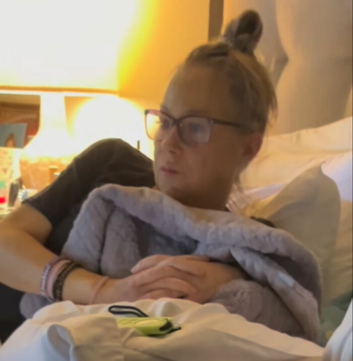 Christina Applegate wearing glasses resting in bed under a blanket.