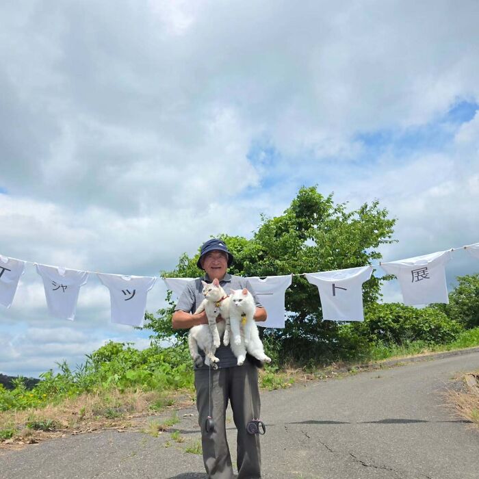 Japanese grandpa smiling outdoor photoshoot holding his two white cats in front of hanging shirts.