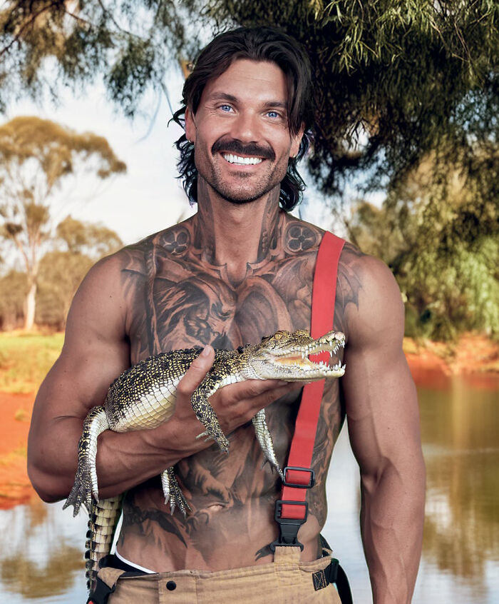 Muscular Australian firefighter with tattoos holding a young crocodile by water