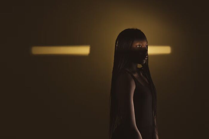 A young woman standing in low light with a beam of light behind her, highlighting human expression in portrait photos.