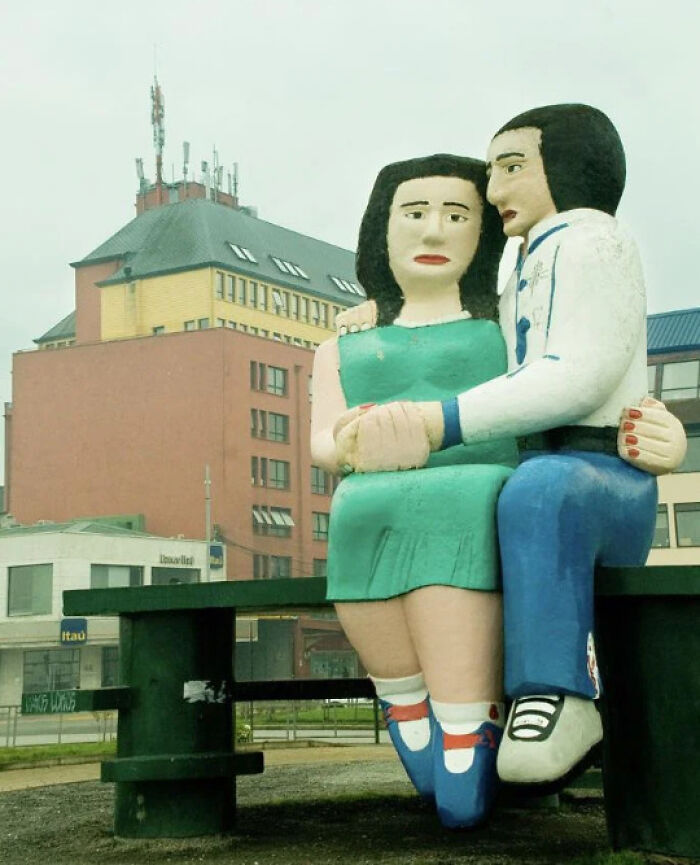 A quirky public art statue of a couple sitting on a bench in front of a city building