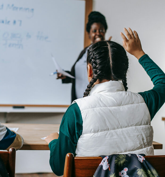 Student raising hand to answer question in classroom with teacher in background