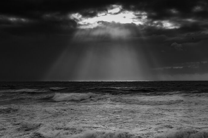 Dramatic sunbeams breaking through dark clouds over stormy ocean waves near black sand in Iceland.