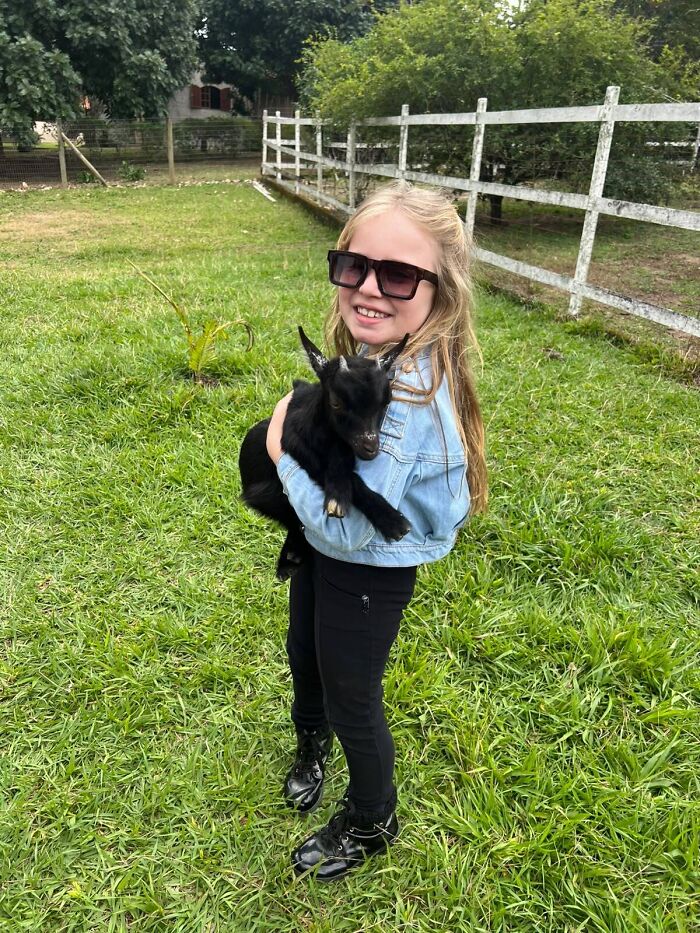 A young girl in sunglasses holding a small black goat outdoors