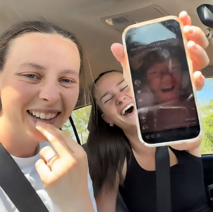 Two women smiling in a car while holding up a phone showing a social media video