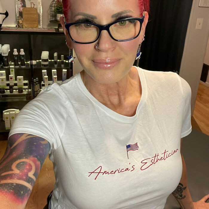 58 years old MAGA influencer wearing America's Esthetician t-shirt with red hair and glasses