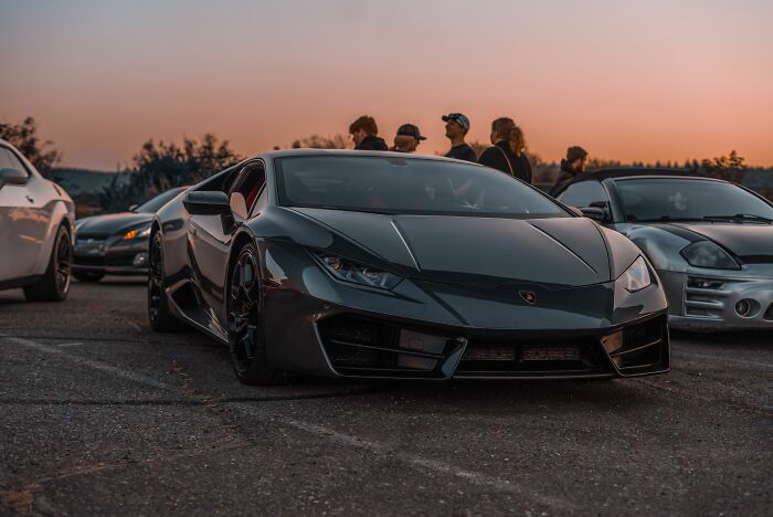 A dark grey luxury sports car parked outdoors surrounded by people at sunset with secrets shared