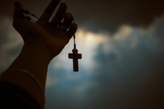 Hand holding cross necklace against cloudy sky symbolizing faith transition