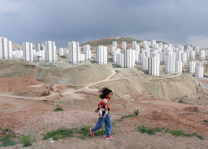 Child holding doll walking near new city buildings, best pics Siena International Photo Awards 2026