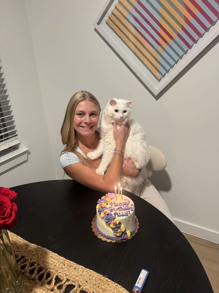 A woman holding a white cat next to a birthday cake, from the viral TikTok trend of sharing life stories with pets
