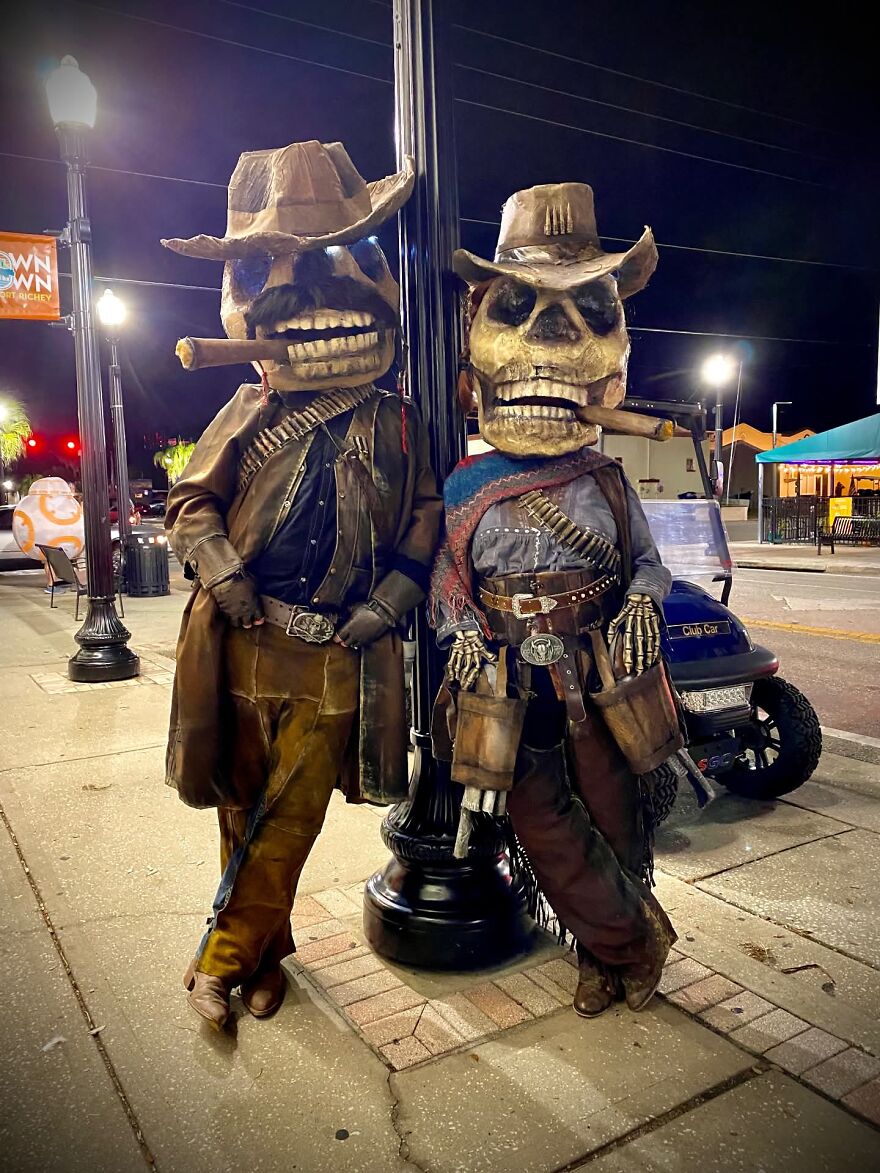 Two people posing outdoors in elaborate skeleton cowboy Halloween costumes with oversized skull heads.
