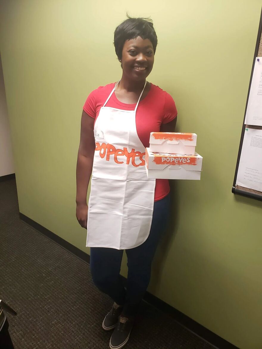 Woman smiling in a Popeyes employee Halloween costume holding chicken boxes and wearing an apron.