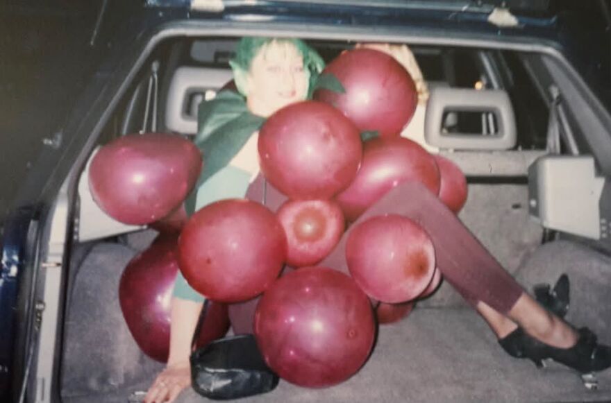 A person dressed as a bunch of purple grapes sitting in the trunk of a car as a fun Halloween costume.