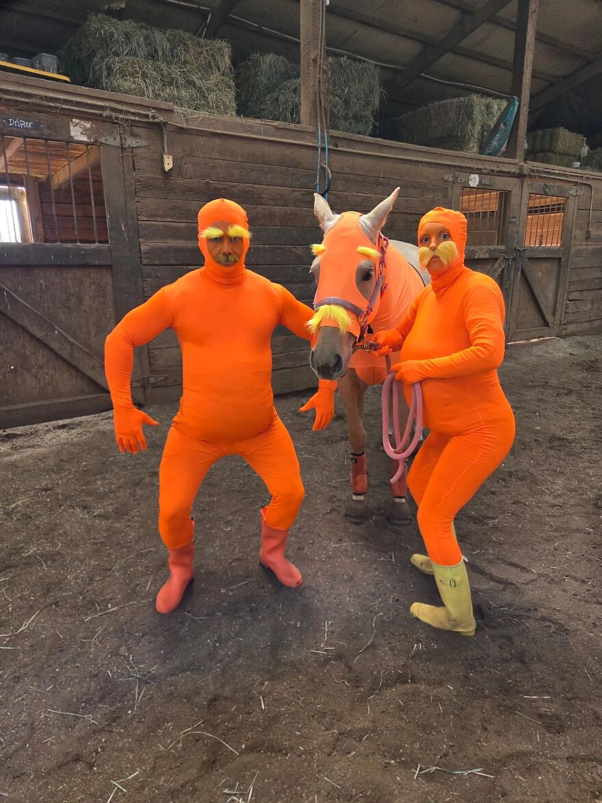 Two people and a horse wearing matching orange Lorax suits inside a barn as creative Halloween costumes.