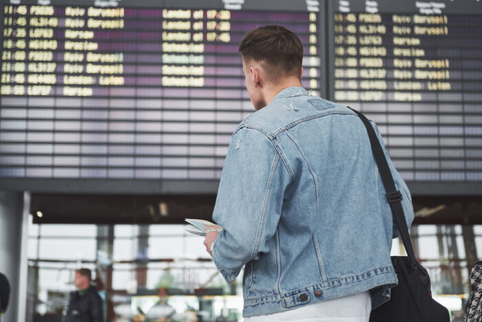 A man in a denim jacket checking a flight tracker board at an airport terminal before taking a flight to Italy.