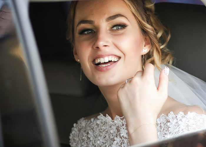 A smiling bride wearing a white lace dress looks out of a car window