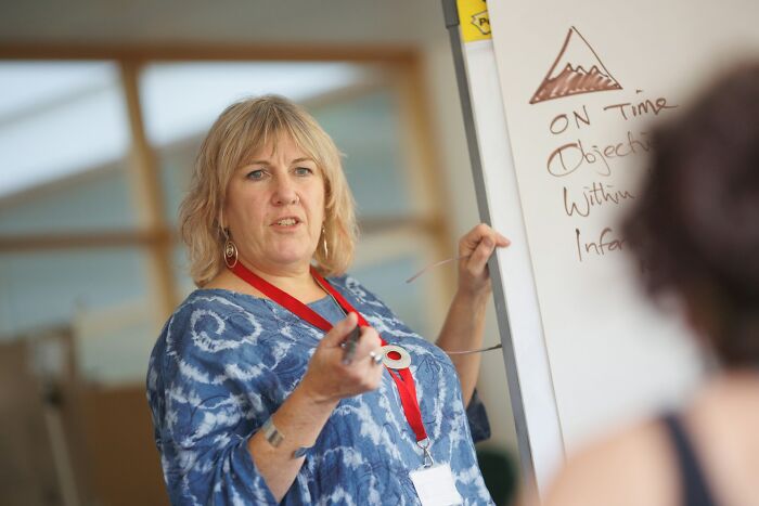 An infuriating teacher gesturing with a pen while explaining a concept on a whiteboard in front of class.