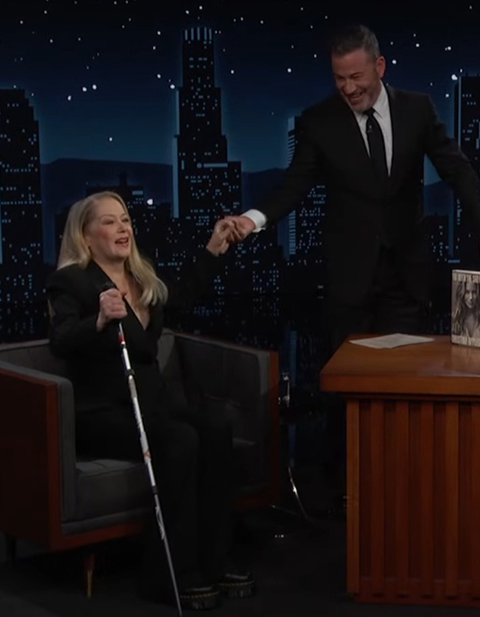 Christina Applegate holding a walking cane while seated next to Jimmy Kimmel on a late night TV show set. Christina Applegate holding a walking cane while seated next to Jimmy Kimmel on a late night TV show set.