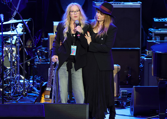 Christina Applegate holding a cane and microphone on stage while supported by another woman. Christina Applegate holding a cane and microphone on stage while supported by another woman.