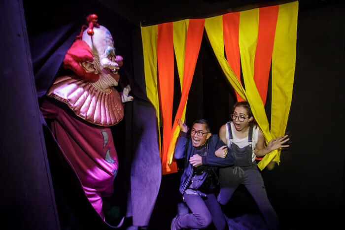 A man and woman scream in fear as a scary clown appears in a haunted house.