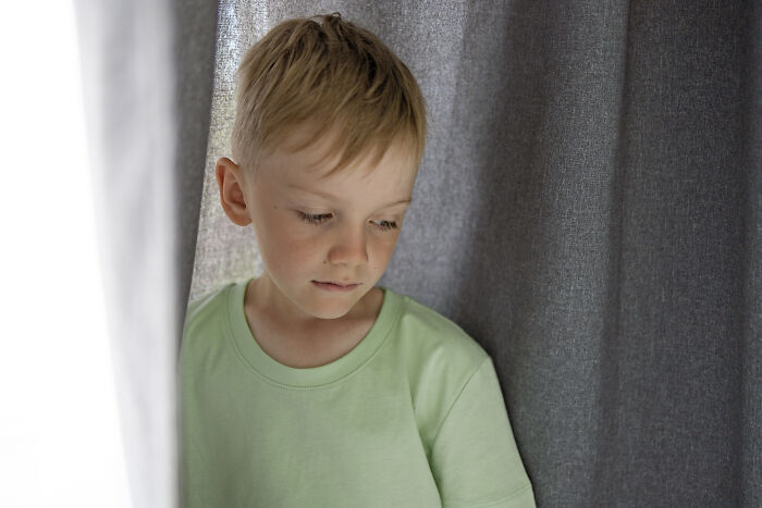 A sad young boy looking down while hiding behind a curtain