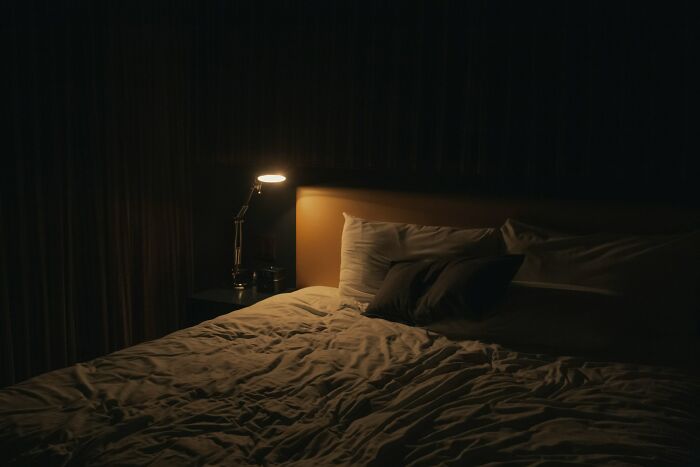 Dimly lit bedroom with crumpled bed and single bedside lamp glowing