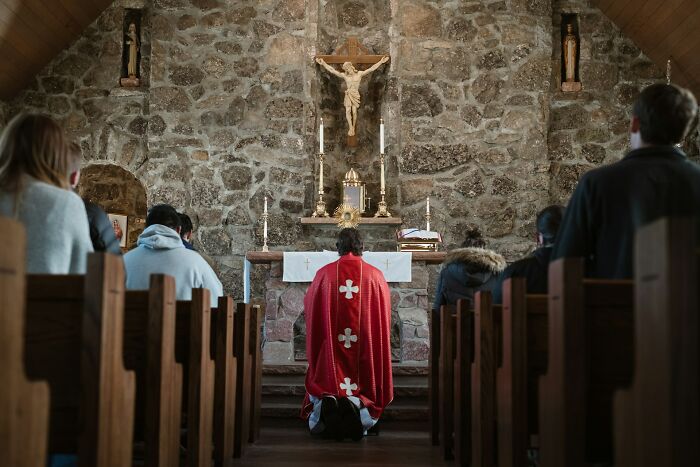 Priest in red robe praying at stone church altar under crucifix statue