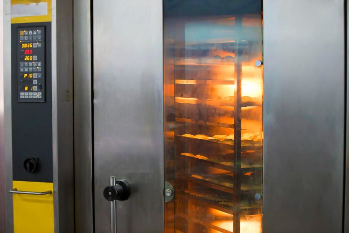 Industrial oven baking multiple trays of bread with visible steam and heat