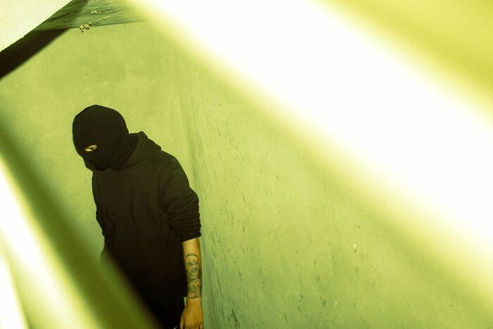 A person in a black balaclava standing in a dimly lit, creepy green hallway during a night shift.
