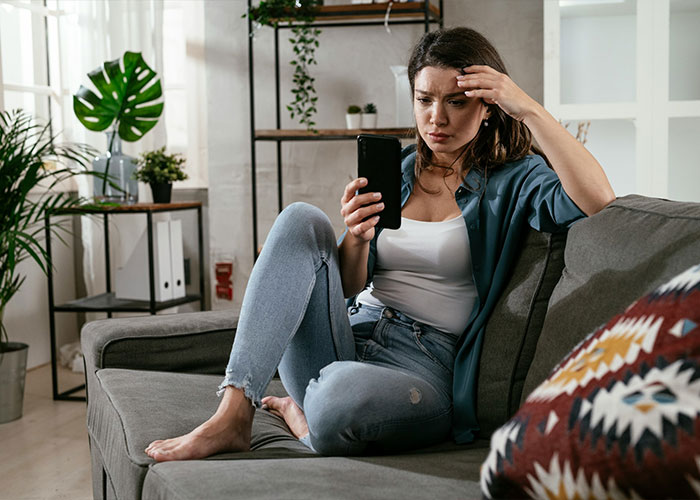 A woman sitting on a couch looking deeply concerned and stressed while looking at her smartphone screen. A woman sitting on a couch looking deeply concerned and stressed while looking at her smartphone screen.