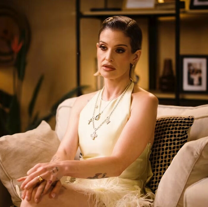 Kelly Osbourne sitting on a couch in a sleeveless top, discussing weight gain and addressing plastic surgery claims.