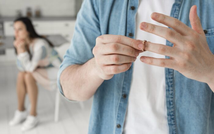 Man taking off wedding ring while contemplating grounds for divorce from his wife in the background.