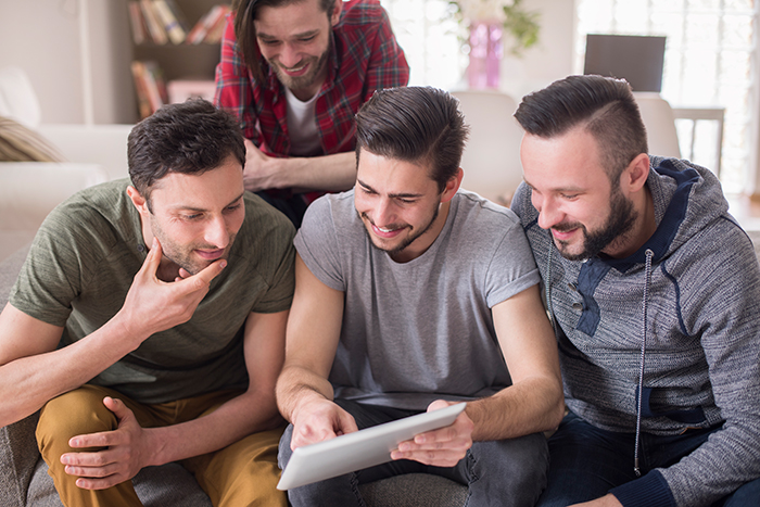 Four young men sitting on a couch smiling and looking at a tablet, representing dad-shamed daughter Four young men sitting on a couch smiling and looking at a tablet, representing dad-shamed daughter