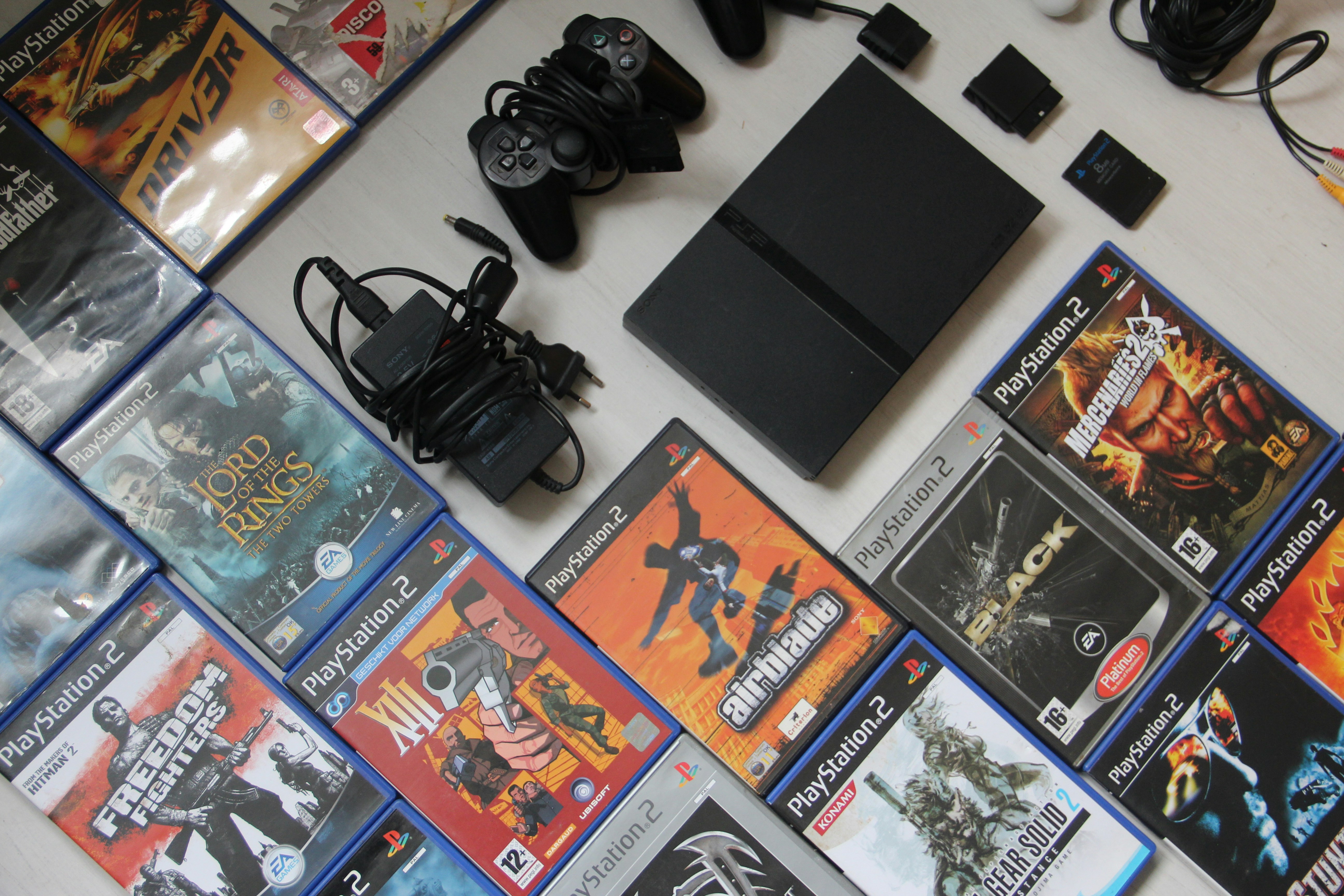 A black PS2 console and controllers surrounded by various video game cases. A black PS2 console and controllers surrounded by various video game cases.