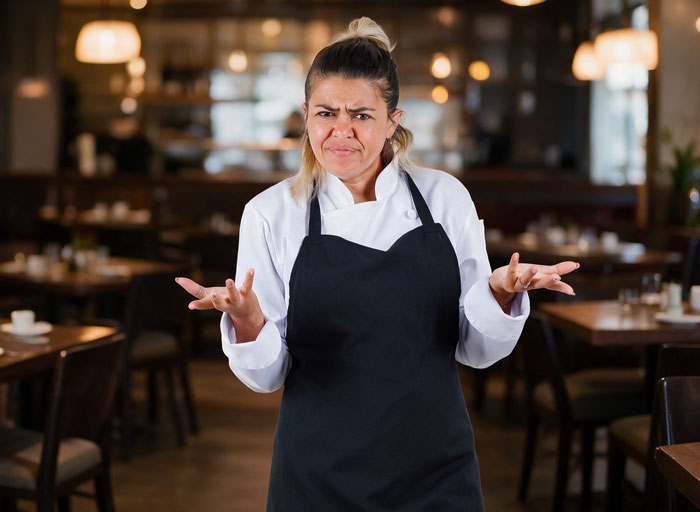 Confused female restaurant worker in a white chef coat and black apron, representing a bossy coworker. Confused female restaurant worker in a white chef coat and black apron, representing a bossy coworker.