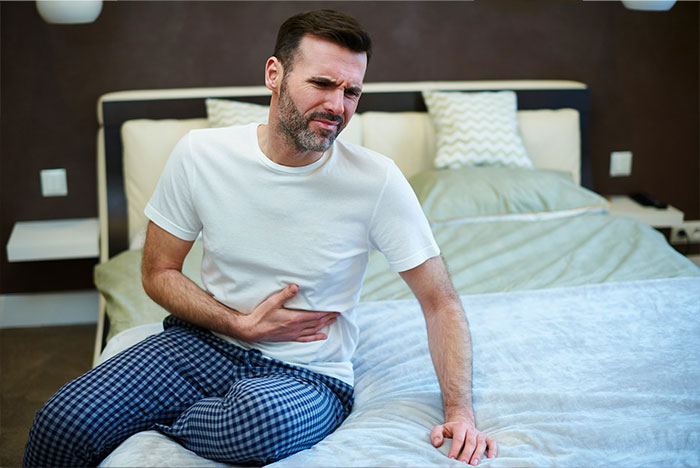 A man sitting on the edge of a bed holding his side in stomach pain. A man sitting on the edge of a bed holding his side in stomach pain.