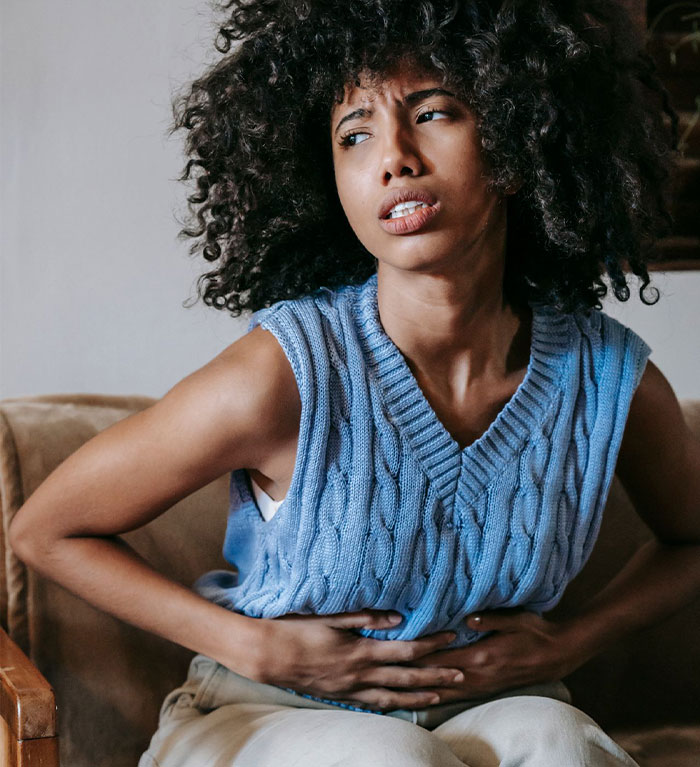 A young woman sitting on a couch holding her stomach in pain or discomfort. A young woman sitting on a couch holding her stomach in pain or discomfort.