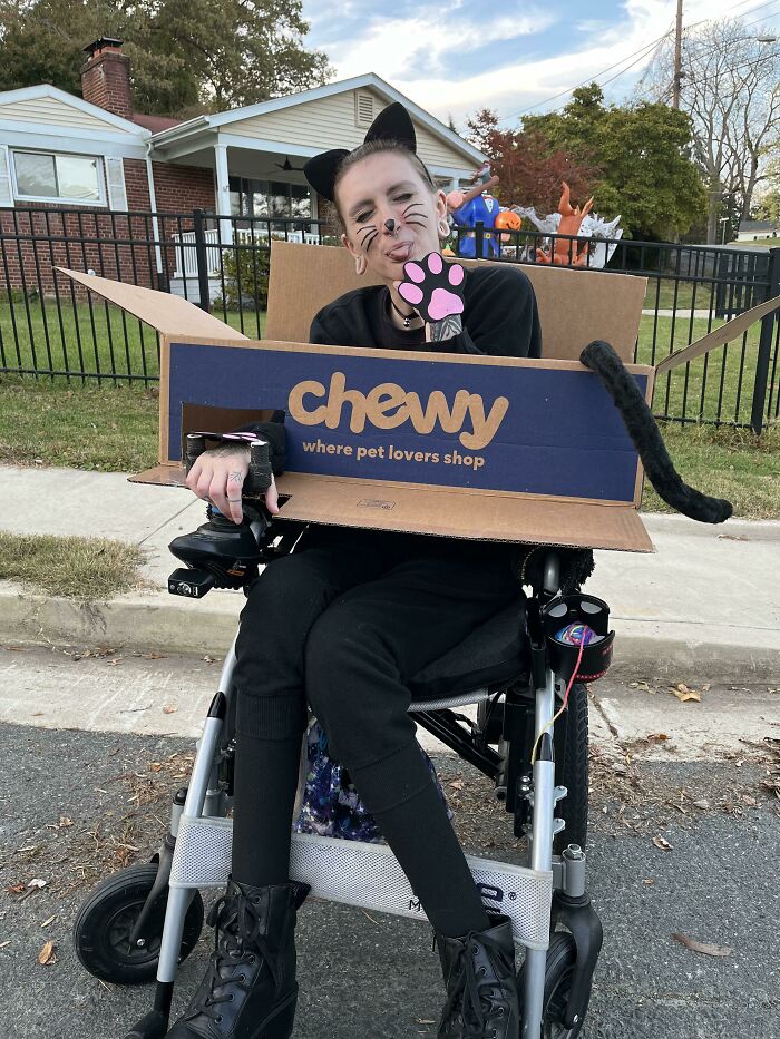 A person in a black cat costume sitting in a wheelchair styled like a cardboard Chewy box for Halloween.
