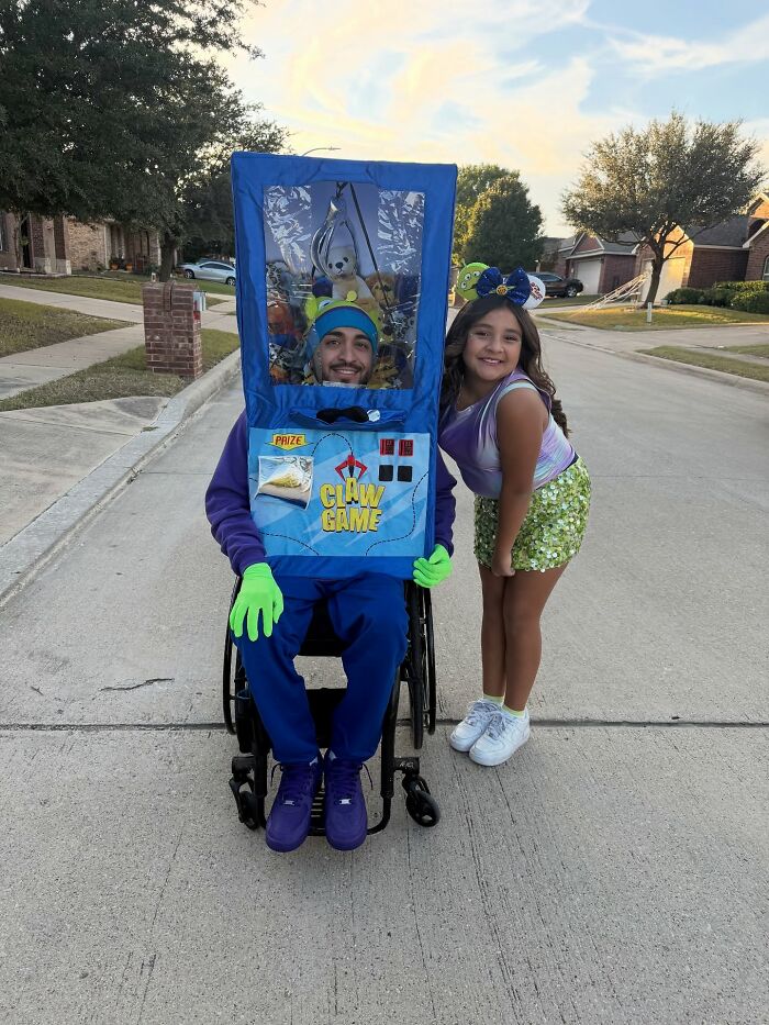A man in a wheelchair formatted as a claw game machine poses with a girl for Halloween.
