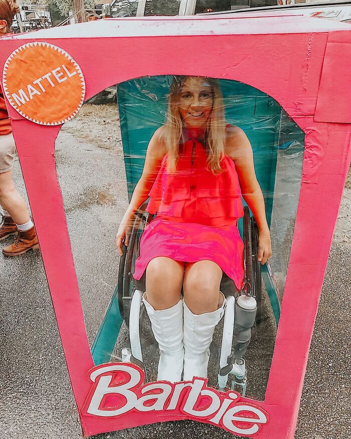 A smiling woman sitting in a wheelchair styled as a pink Barbie doll box for Halloween.